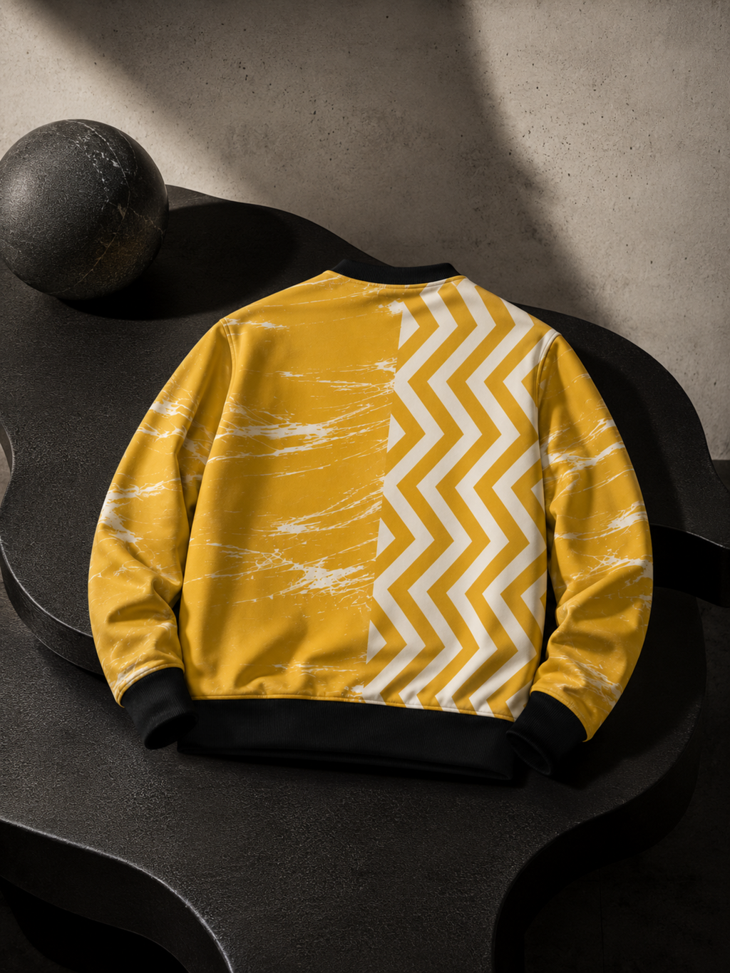 A yellow sweatshirt with a white chevron pattern on one side and a white marble-like print on the other, displayed on a dark sculptural surface.