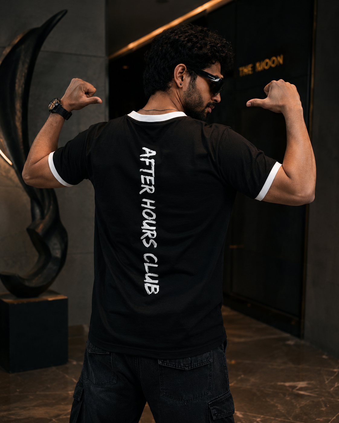 Man wearing a black t-shirt with 'After Hours Club' text on the back, flexing his muscles in a dimly lit room.