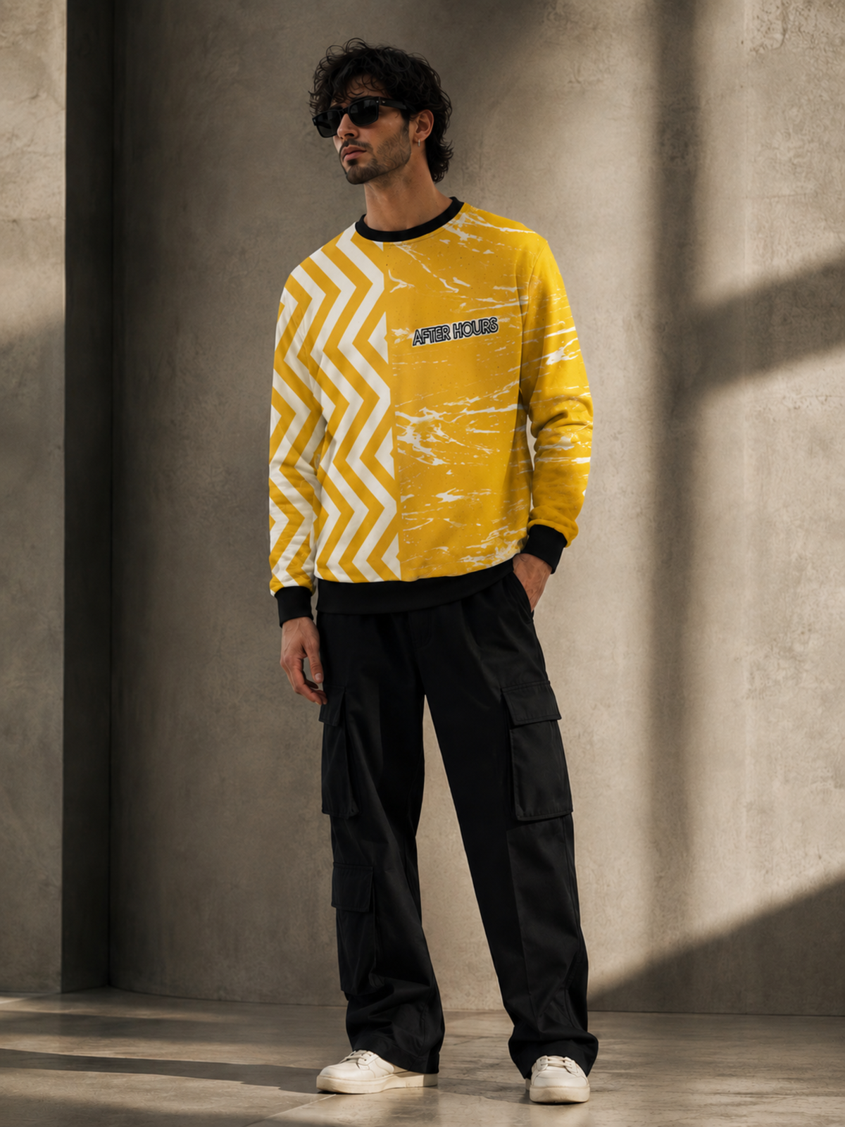 A man wearing a yellow and white patterned long-sleeve sweatshirt, black cargo pants, and white sneakers, standing in front of a textured concrete wall.