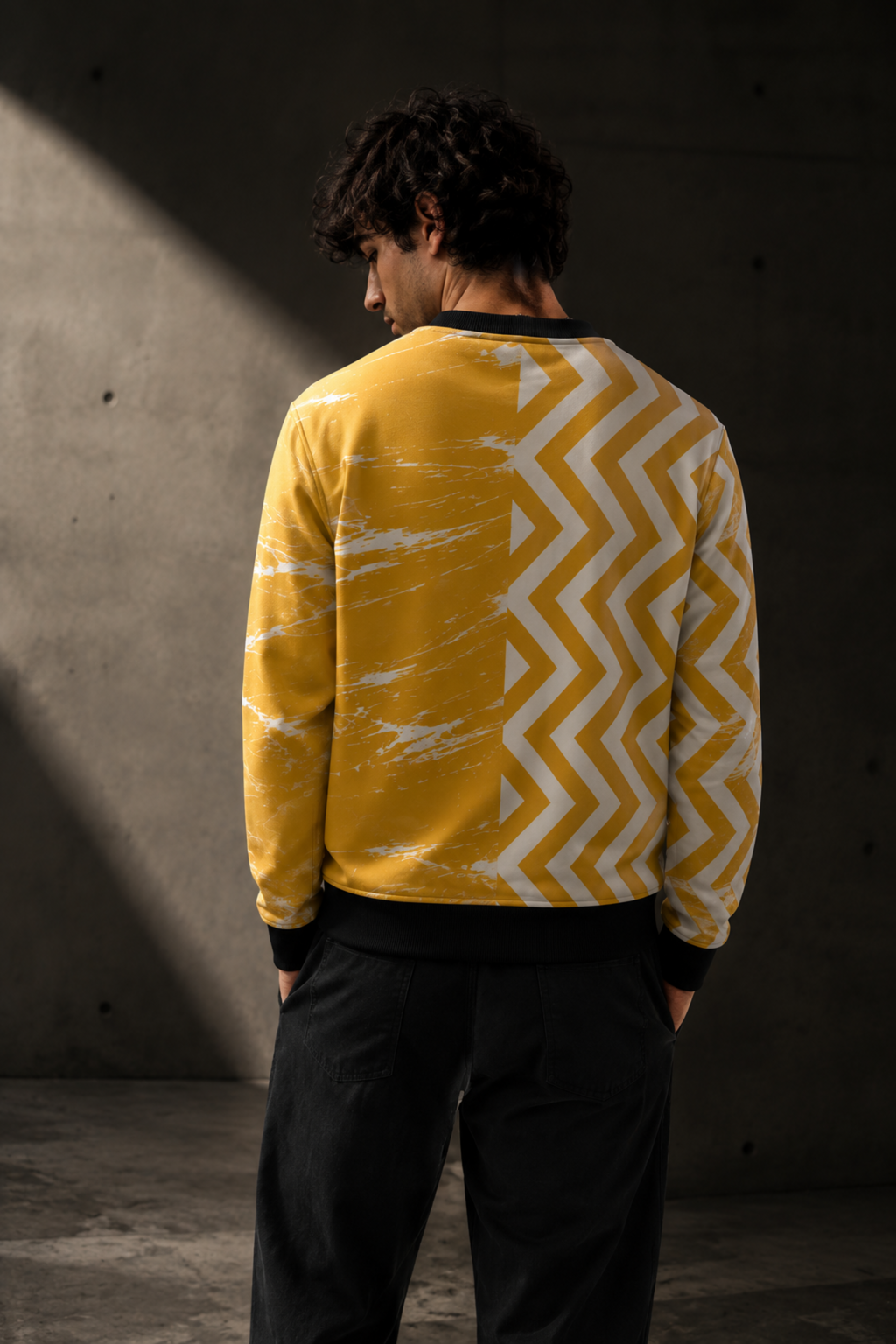 A man viewed from behind wearing a yellow crewneck sweatshirt with a white chevron pattern on one side and a white marble-like pattern on the other, paired with black pants, standing against a dark concrete wall.