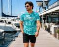 A man wearing a turquoise and white all over print 'AFTER HOURS' printed on the chest, walking on a wooden dock next to a marina with boats and a modern building.