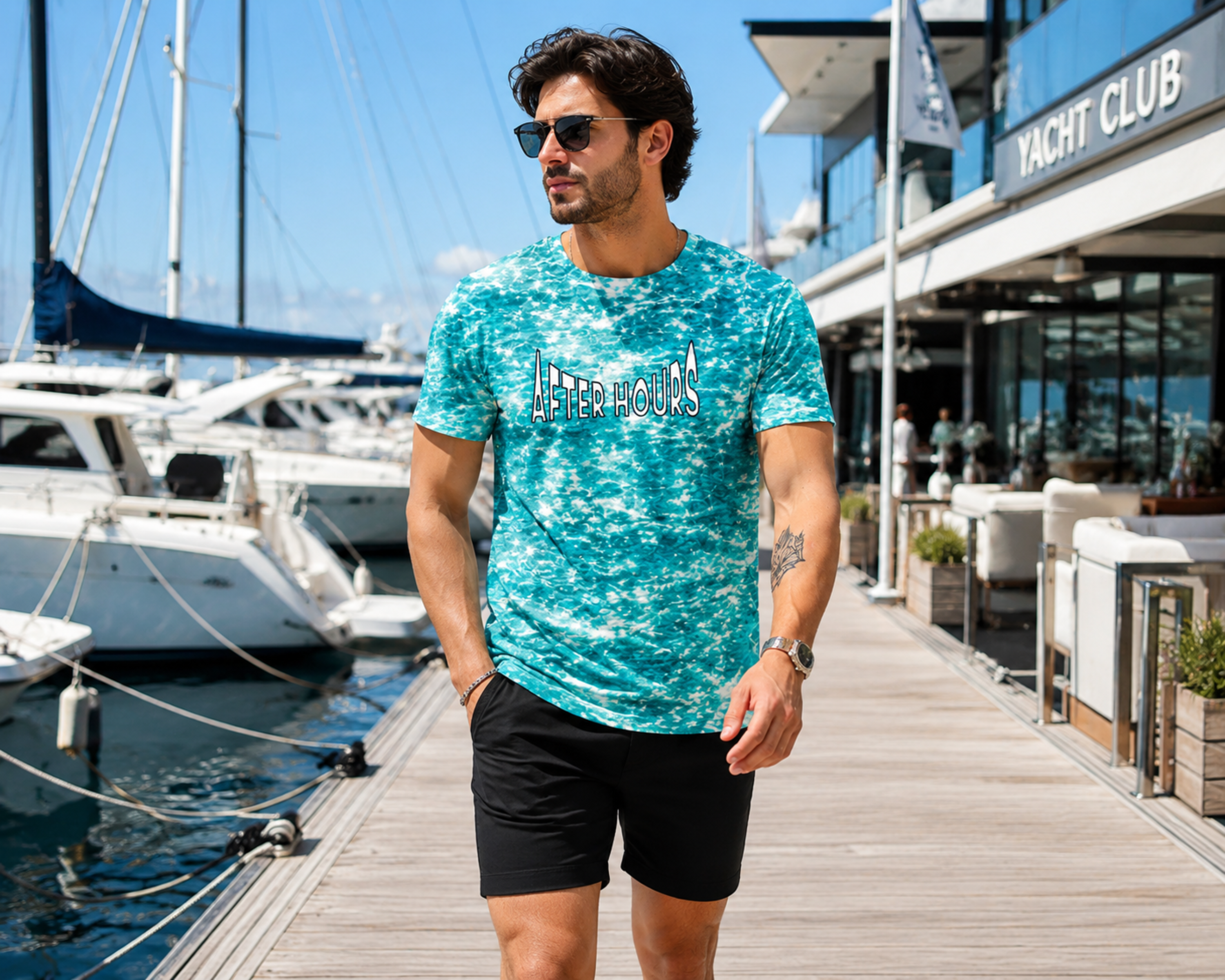 A man wearing a turquoise and white all over print 'AFTER HOURS' printed on the chest, walking on a wooden dock next to a marina with boats and a modern building.