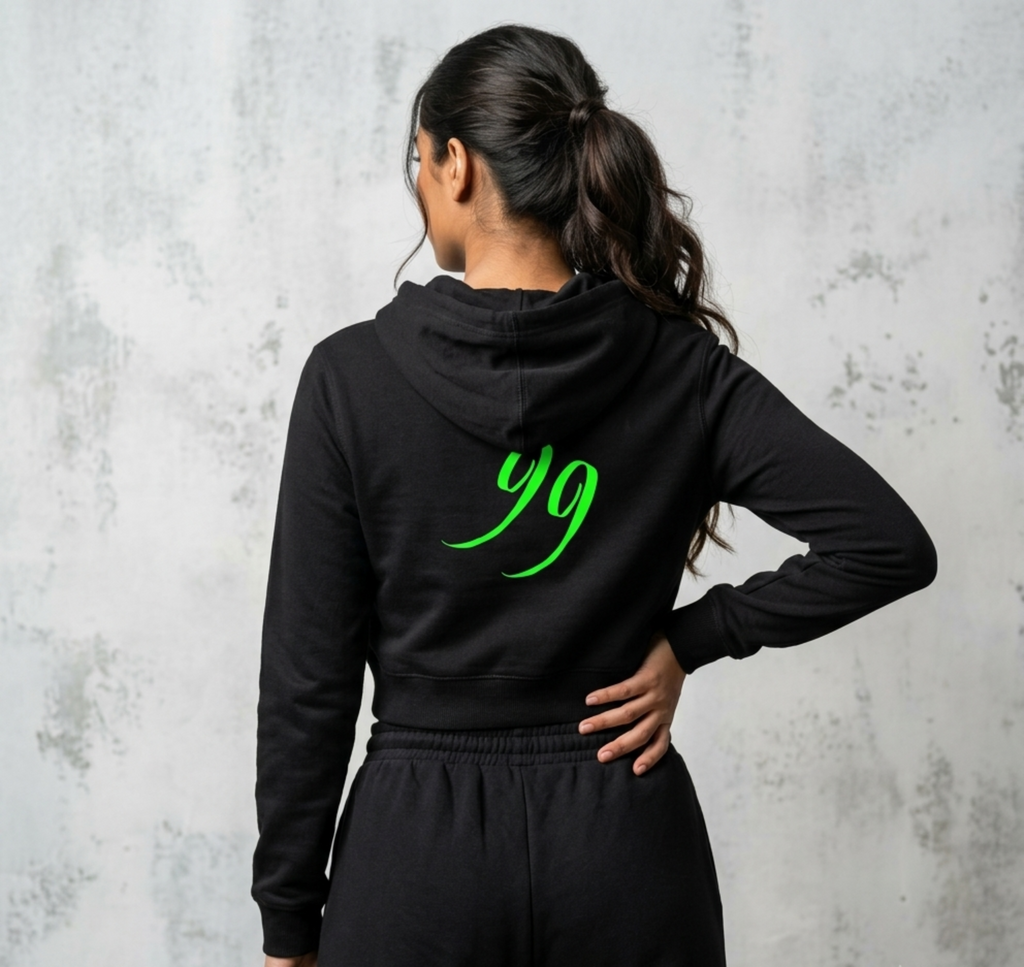 A woman shown from the back wearing a black cropped hoodie with a bright green '99' graphic on the back, paired with matching black sweatpants, set against a textured grey wall.