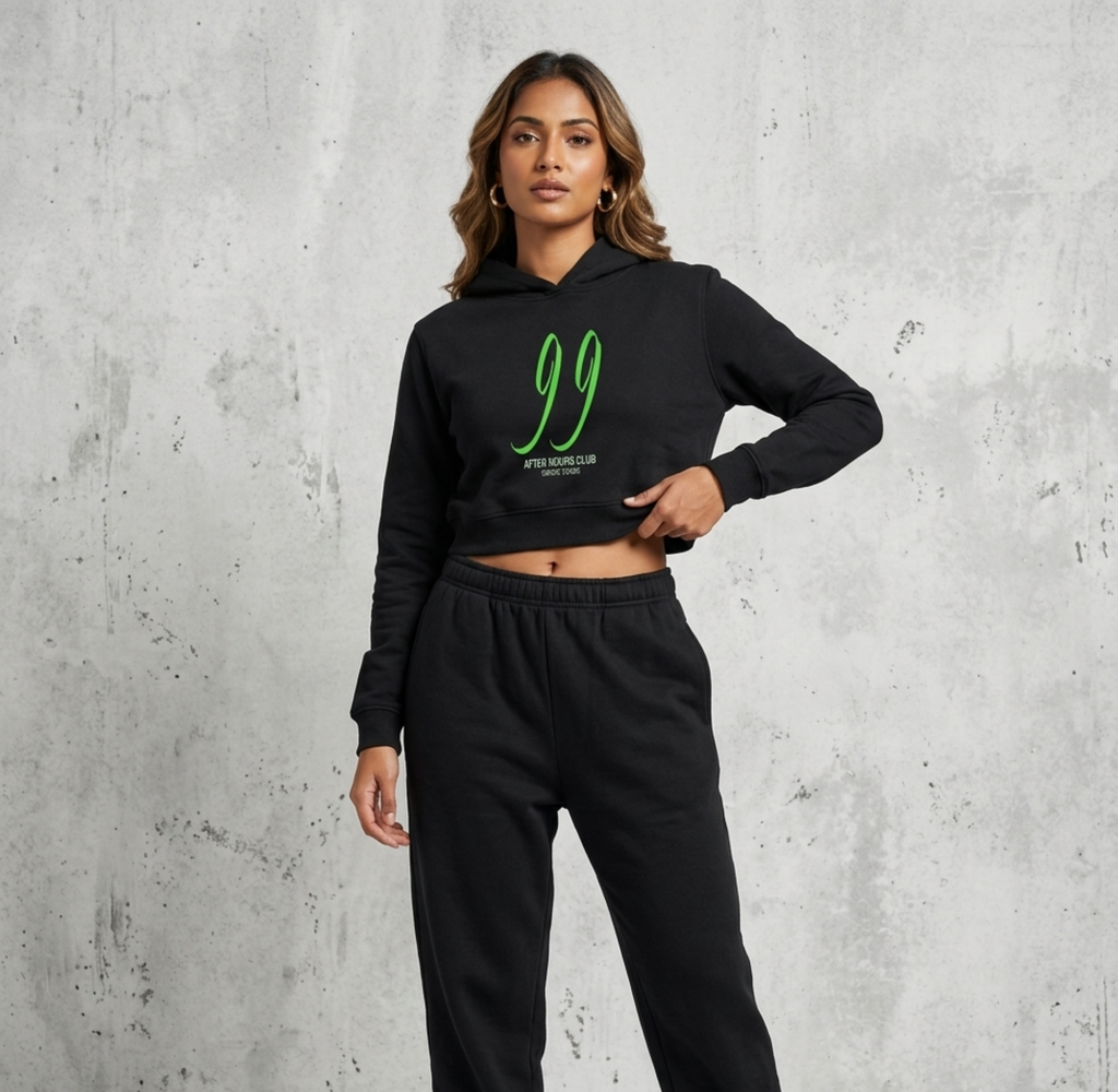 A woman modeling a black cropped hoodie and matching sweatpants set against a textured gray concrete wall. The hoodie features a bright green '99' graphic with 'AFTER HOURS CLUB SINCE 2025' printed below it.