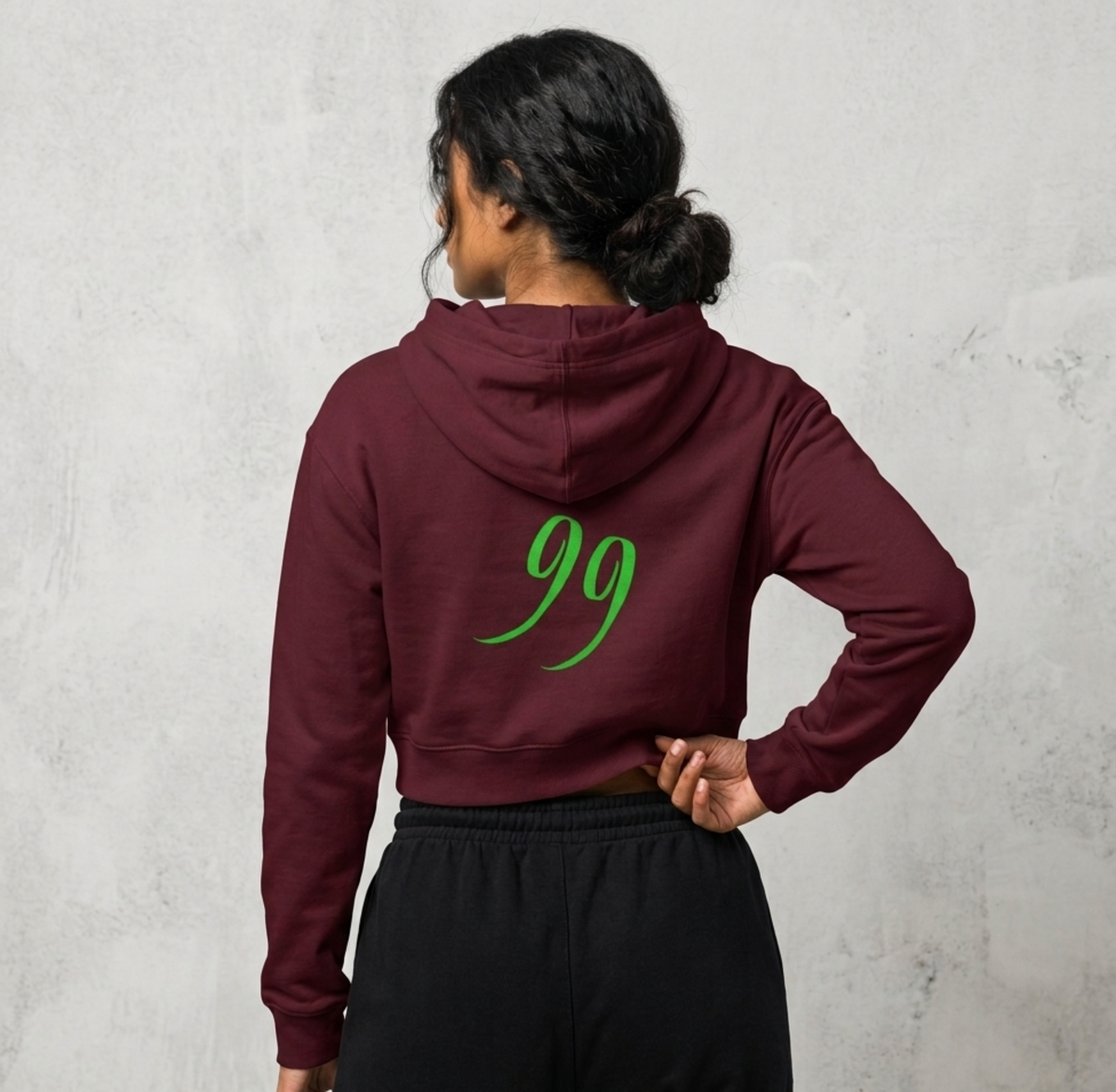 A woman shown from the back wearing a cropped maroon hoodie with a bright green '99' graphic on the back, paired with black sweatpants, standing against a textured light grey wall.