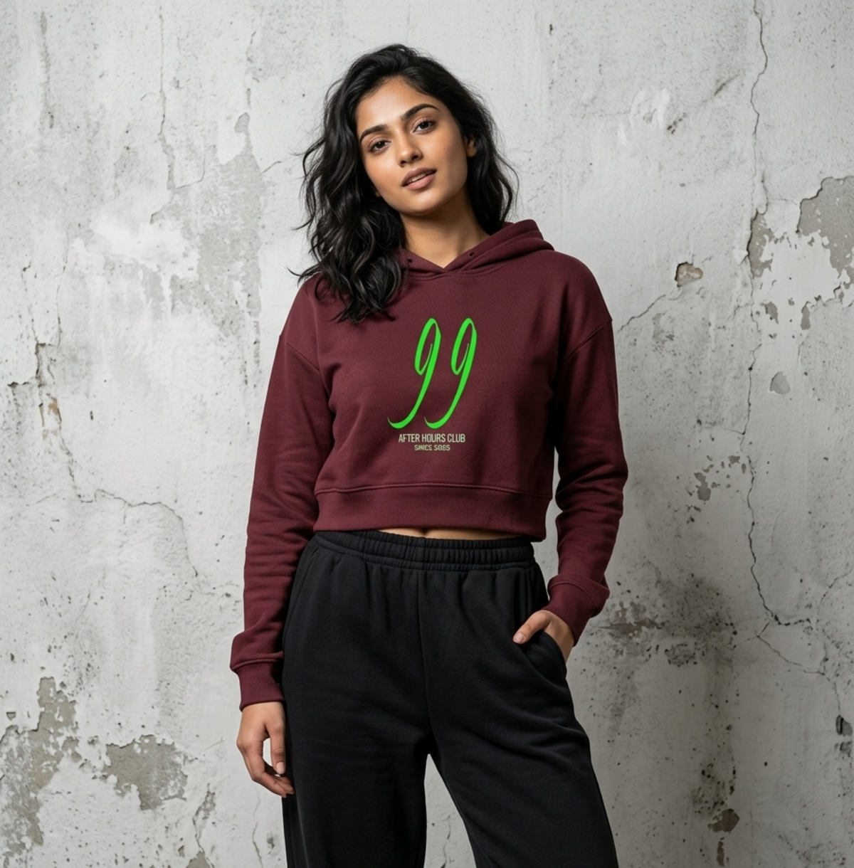 A woman stands against a textured concrete wall wearing a cropped maroon hoodie with a green graphic print and black sweatpants.