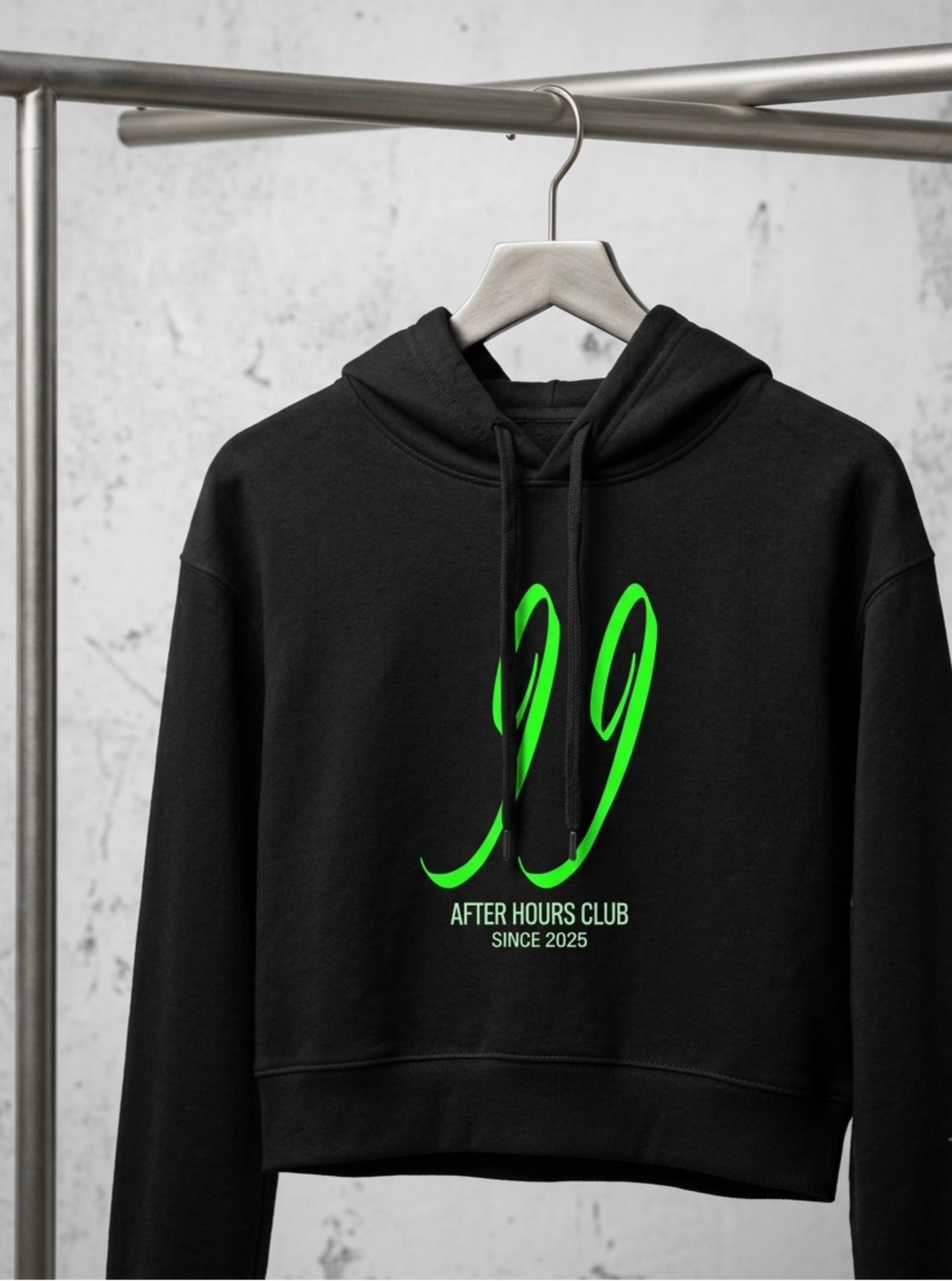 A black cropped hoodie hanging on a wooden hanger from a metal clothing rack, featuring a bright green graphic of the number '99' and the text 'AFTER HOURS CLUB' and 'SINCE 2025' on the front.
