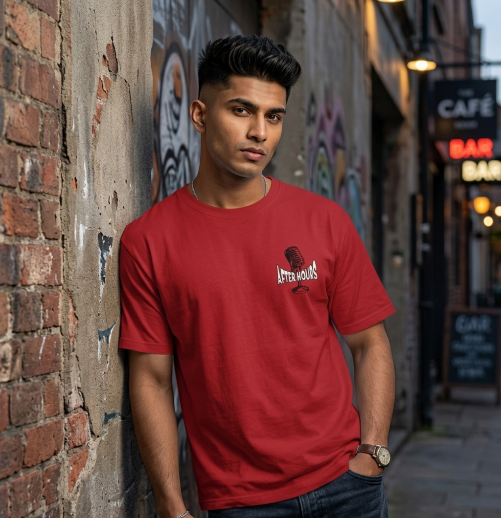 A man wearing a red crewneck t-shirt with a small white graphic on the left chest, leaning against a brick wall in an outdoor alleyway.