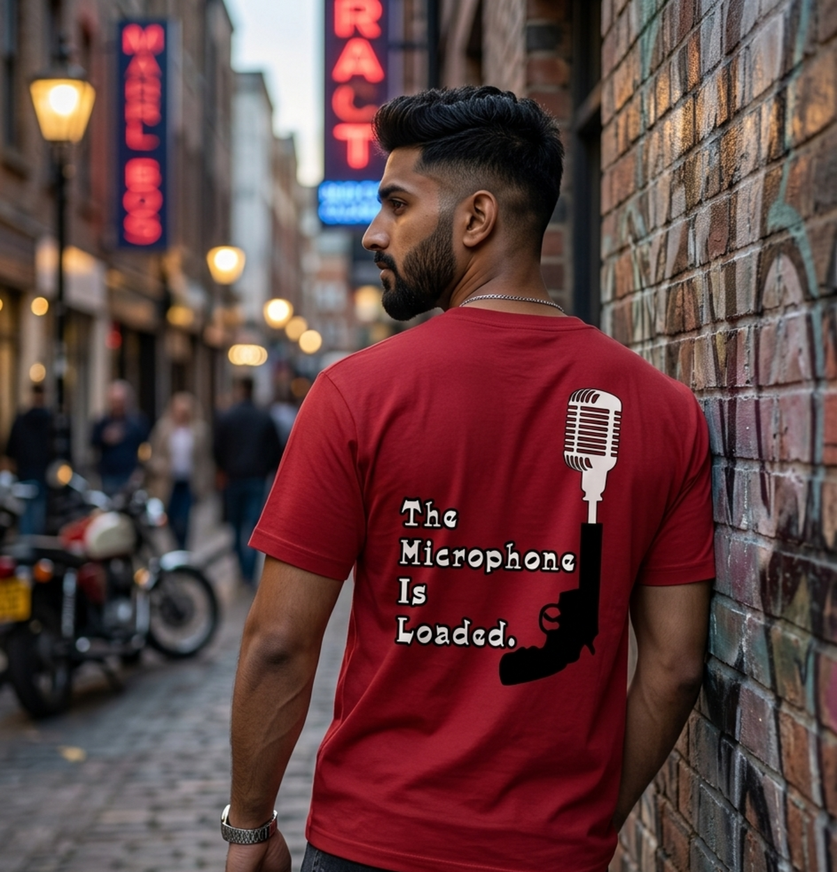 A man standing on a cobblestone street wearing a red t-shirt with a graphic on the back that reads 'The Microphone Is Loaded.' next to a brick wall.