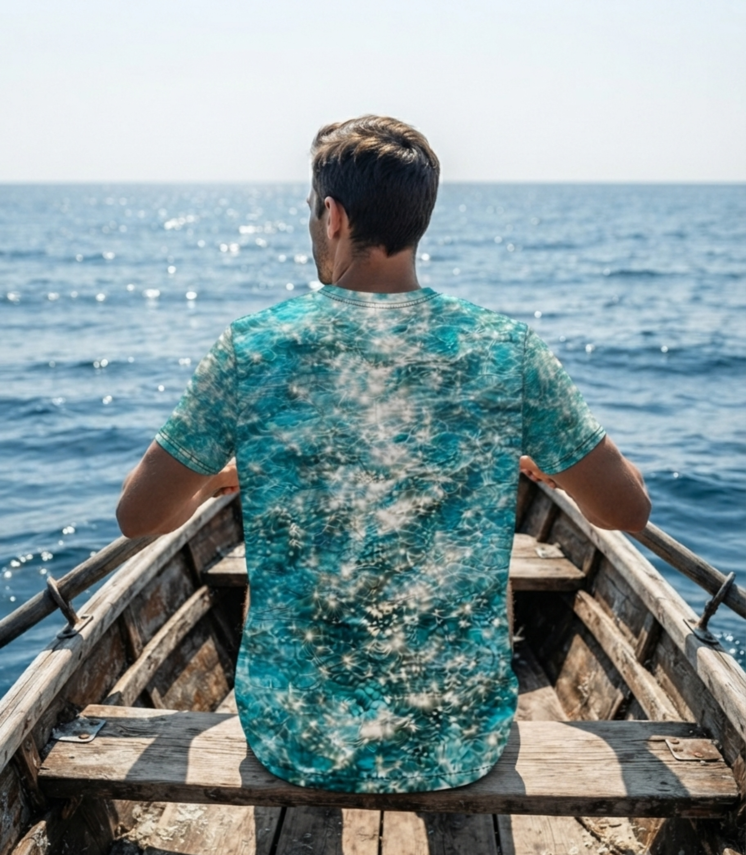 A man sitting in the back of a wooden boat, viewed from behind, wearing a short-sleeve t-shirt with a turquoise and white abstract pattern. The background shows a sunny blue ocean under a pale sky.