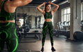 A woman in a gym wearing a matching green and black athletic set with a circular pattern, standing in front of a mirror that reflects her profile. The background shows exercise equipment like treadmills and weight machines.