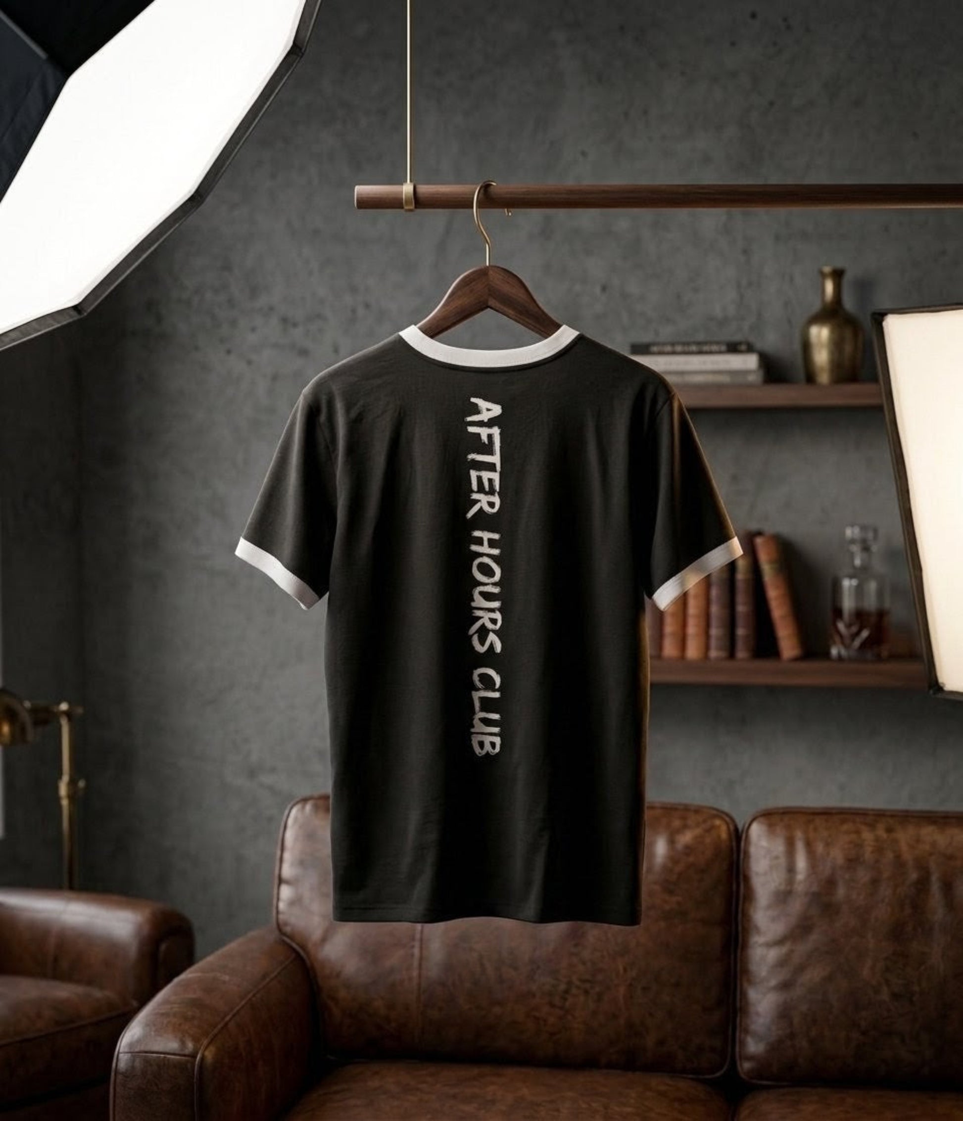 Black t-shirt with 'After Hours Club' text on a hanger against a gray wall with a brown leather couch.