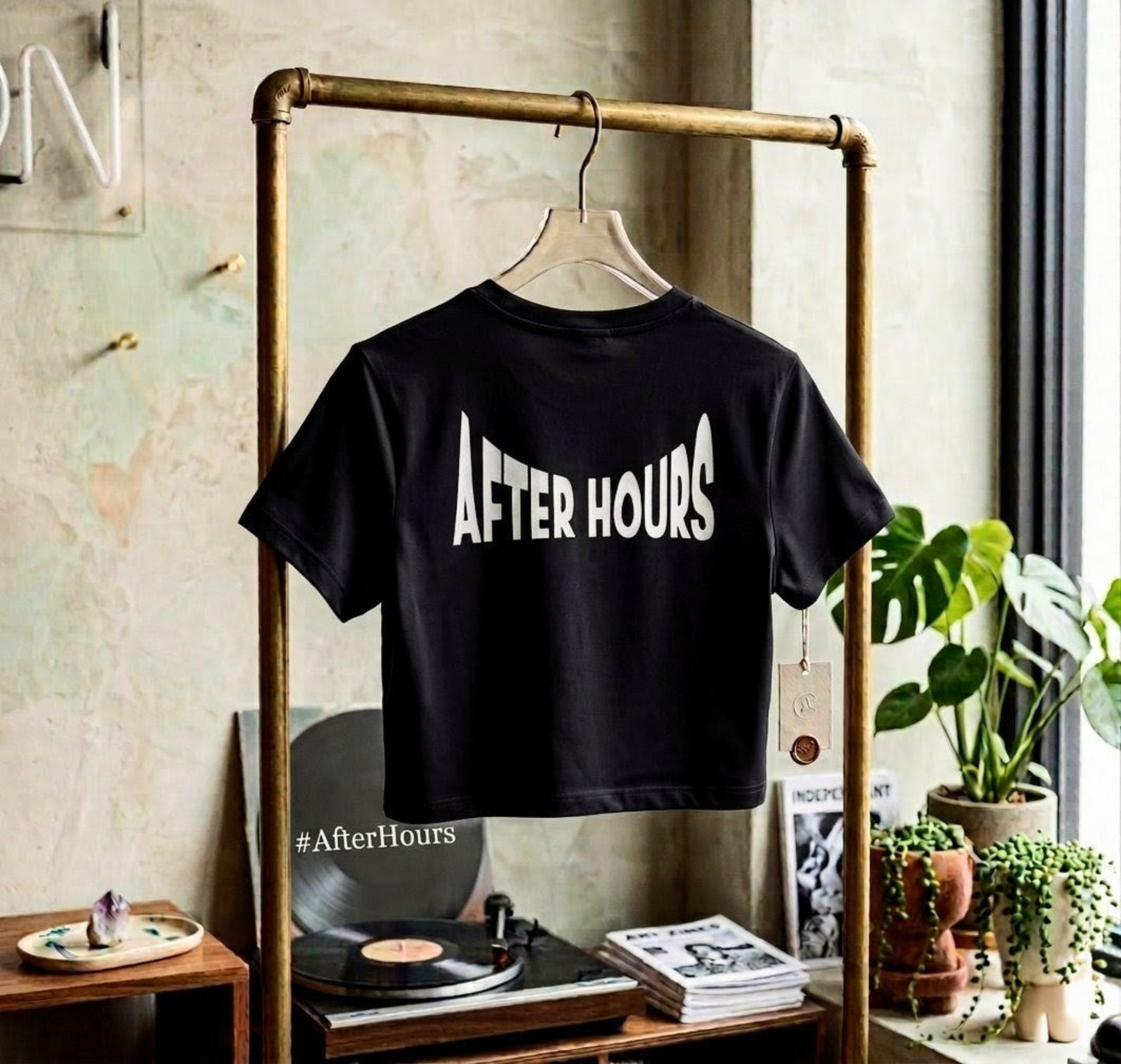 Black t-shirt with 'AFTER HOURS' text hanging on a rack in a room with plants and records.