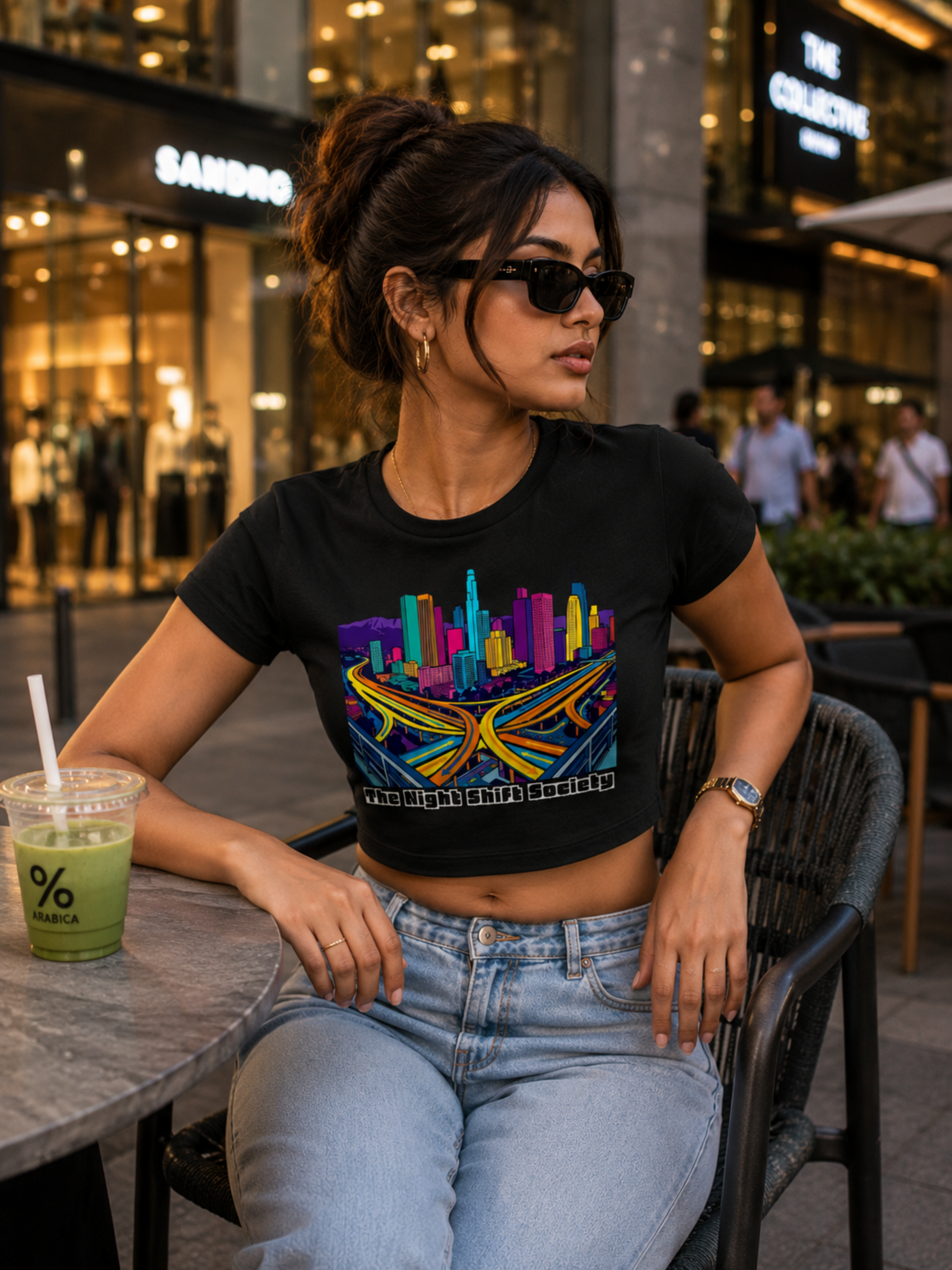 Woman sitting outdoors at night wearing a black t-shirt with a colorful cityscape design.