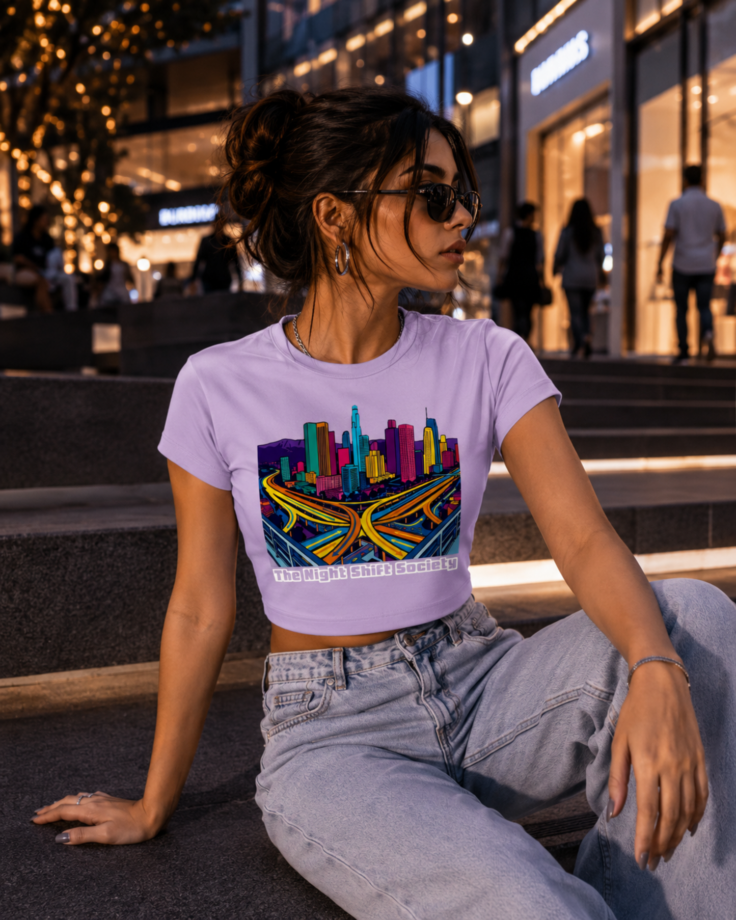 Woman wearing a lavender t-shirt with a colourful cityscape design sitting on steps at night.