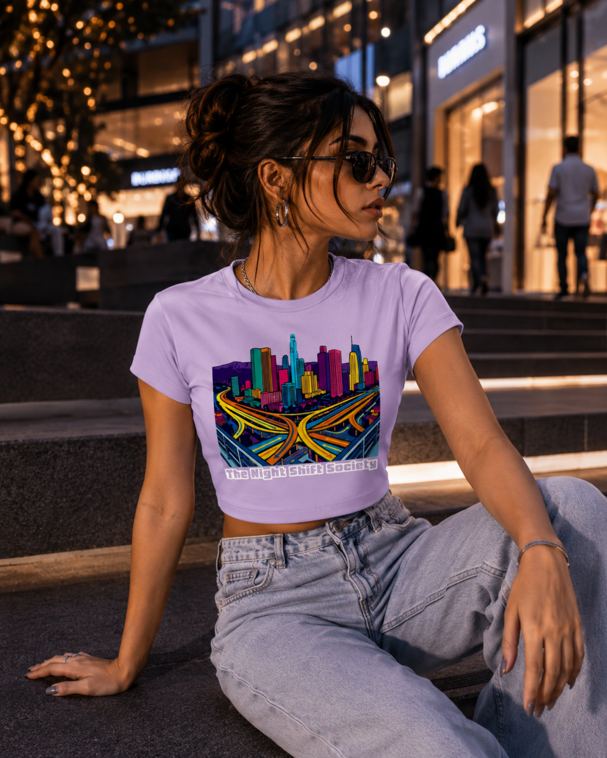 Woman wearing a lavender t-shirt with a colourful cityscape design sitting on steps at night.