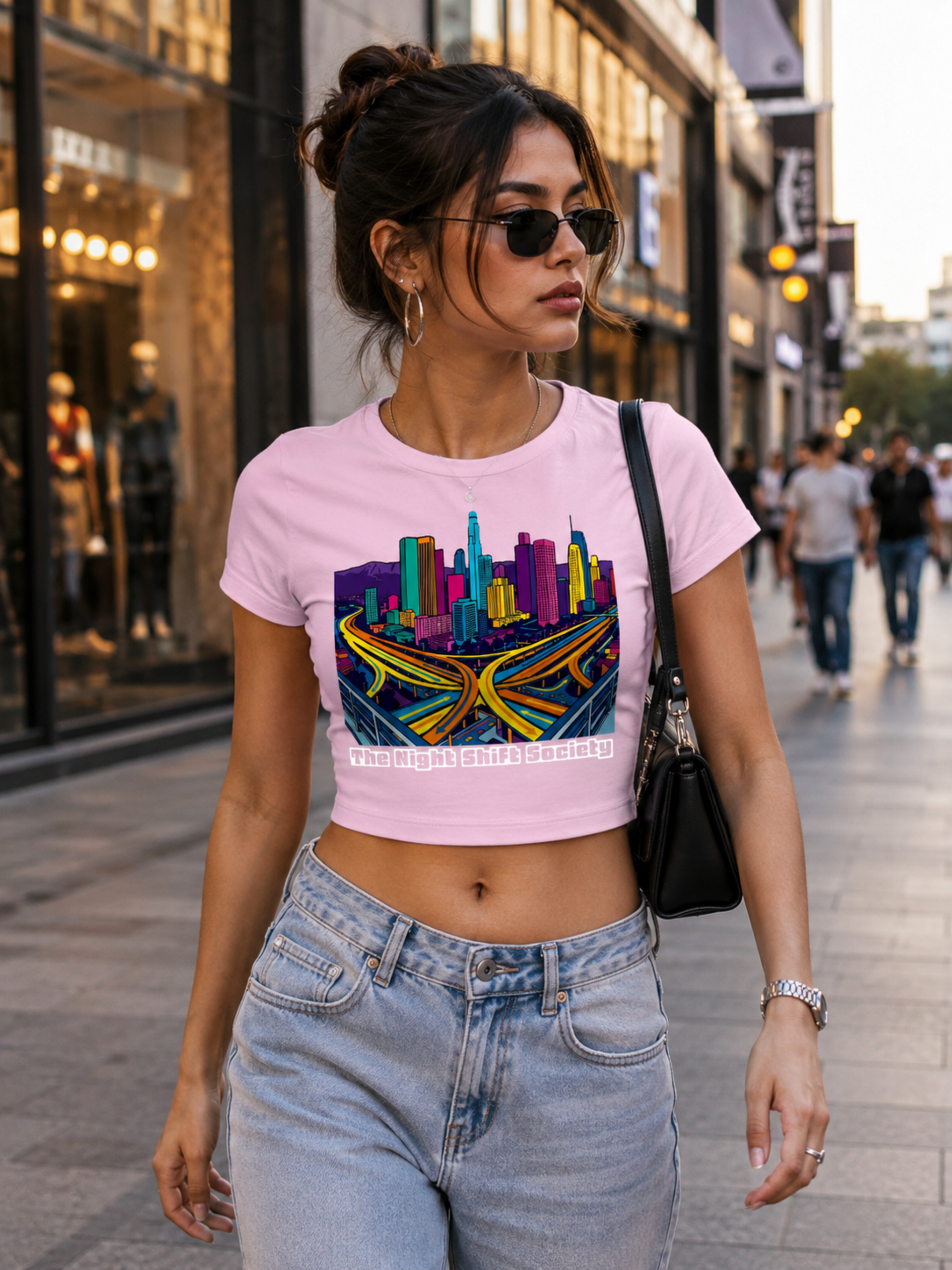 Woman walking on a city street wearing a pink crop top with a colorful graphic design and blue jeans.