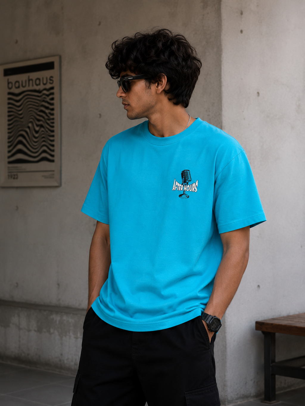 A man wearing a bright sky blue t-shirt with a small graphic on the left chest, paired with black cargo pants, standing against a concrete wall.