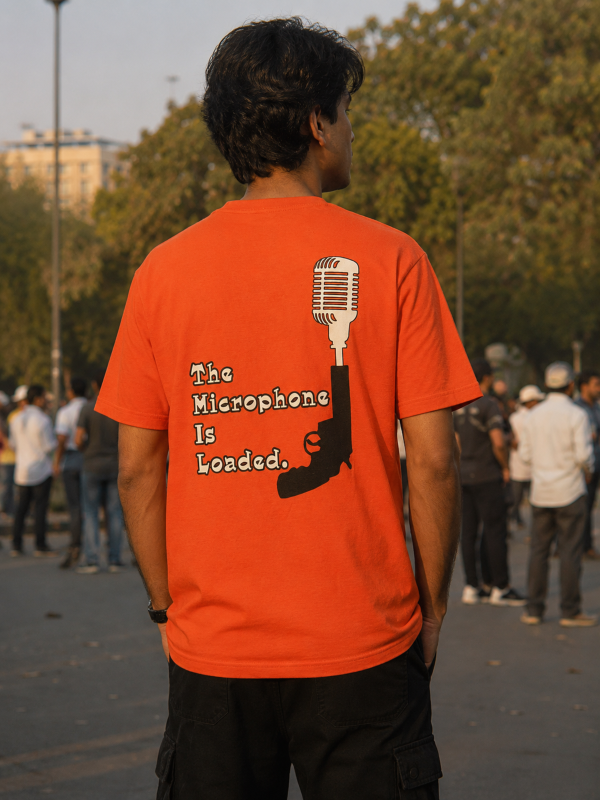 A man viewed from behind wearing a bright orange t-shirt with a graphic on the back featuring a microphone and the text 'The Microphone Is Loaded.' in white and black, set against an outdoor park background.