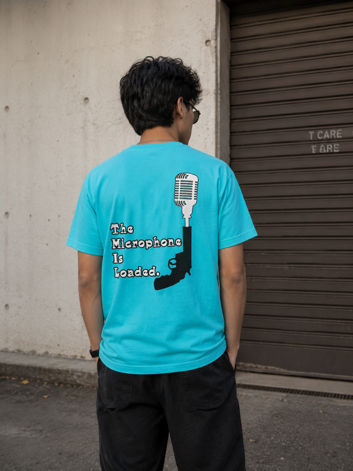 A man viewed from behind wearing a bright sky blue graphic t-shirt with a black and white design that reads 'The Microphone Is Loaded.' next to a handgun silhouette, paired with black pants, standing in front of a concrete wall and a brown metal garage door.