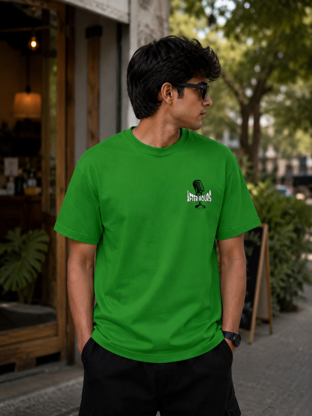 A man wearing a bright green crewneck t-shirt with a small black and white graphic on the left chest, standing outdoors on a sidewalk in front of a storefront.