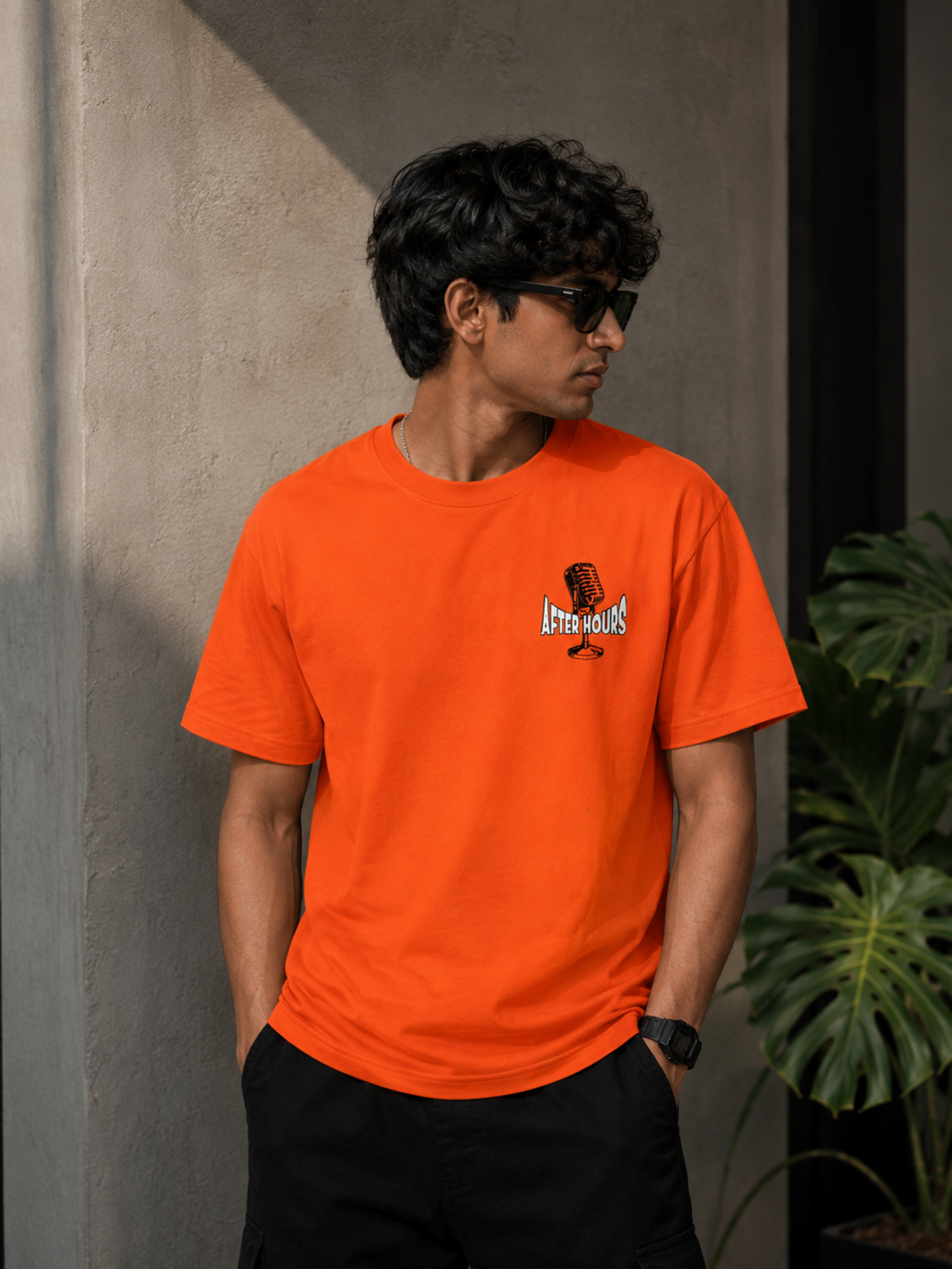 A man wearing a bright orange crewneck t-shirt with a small graphic on the left chest, paired with black pants, standing against a textured concrete wall.