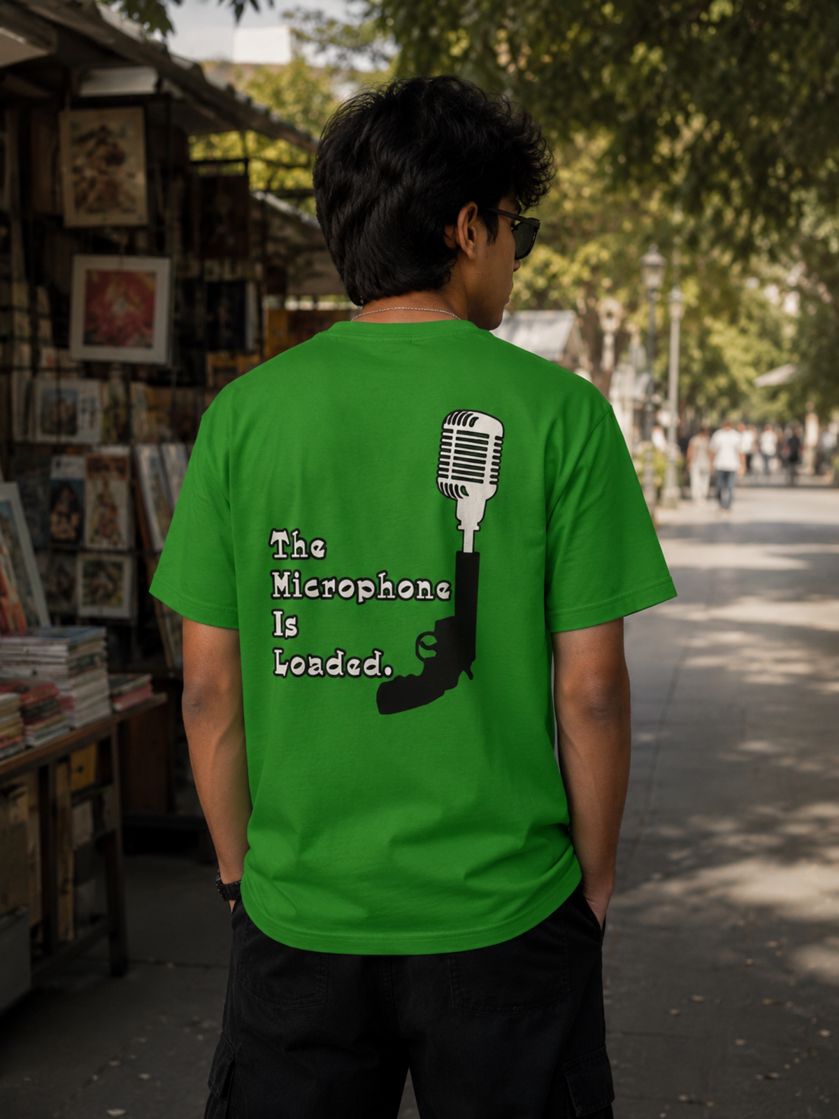 A man viewed from behind wearing a bright green t-shirt with a graphic on the back. The graphic features a vintage microphone and a pistol silhouette with the text 'The Microphone Is Loaded.