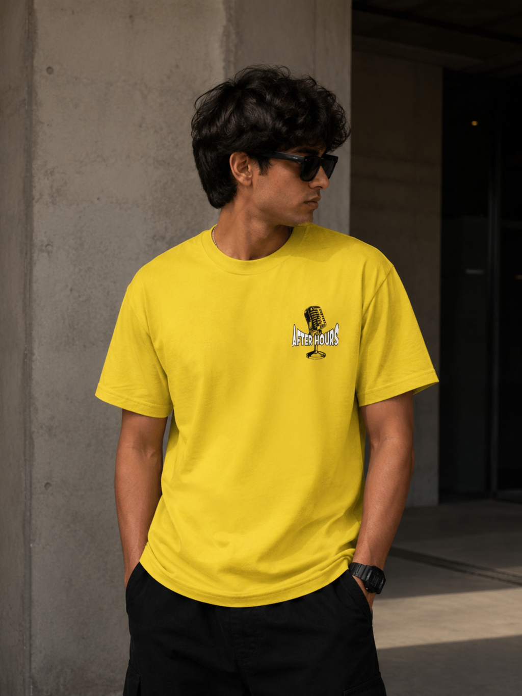 A man wearing a bright yellow crewneck t-shirt with a small graphic on the left chest, paired with black pants and sunglasses, standing against a concrete wall.