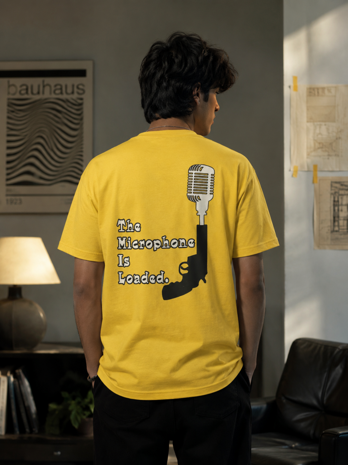 A man viewed from behind wearing a bright yellow t-shirt with a graphic print of a microphone and pistol and the text 'The Microphone Is Loaded.' in a room setting.