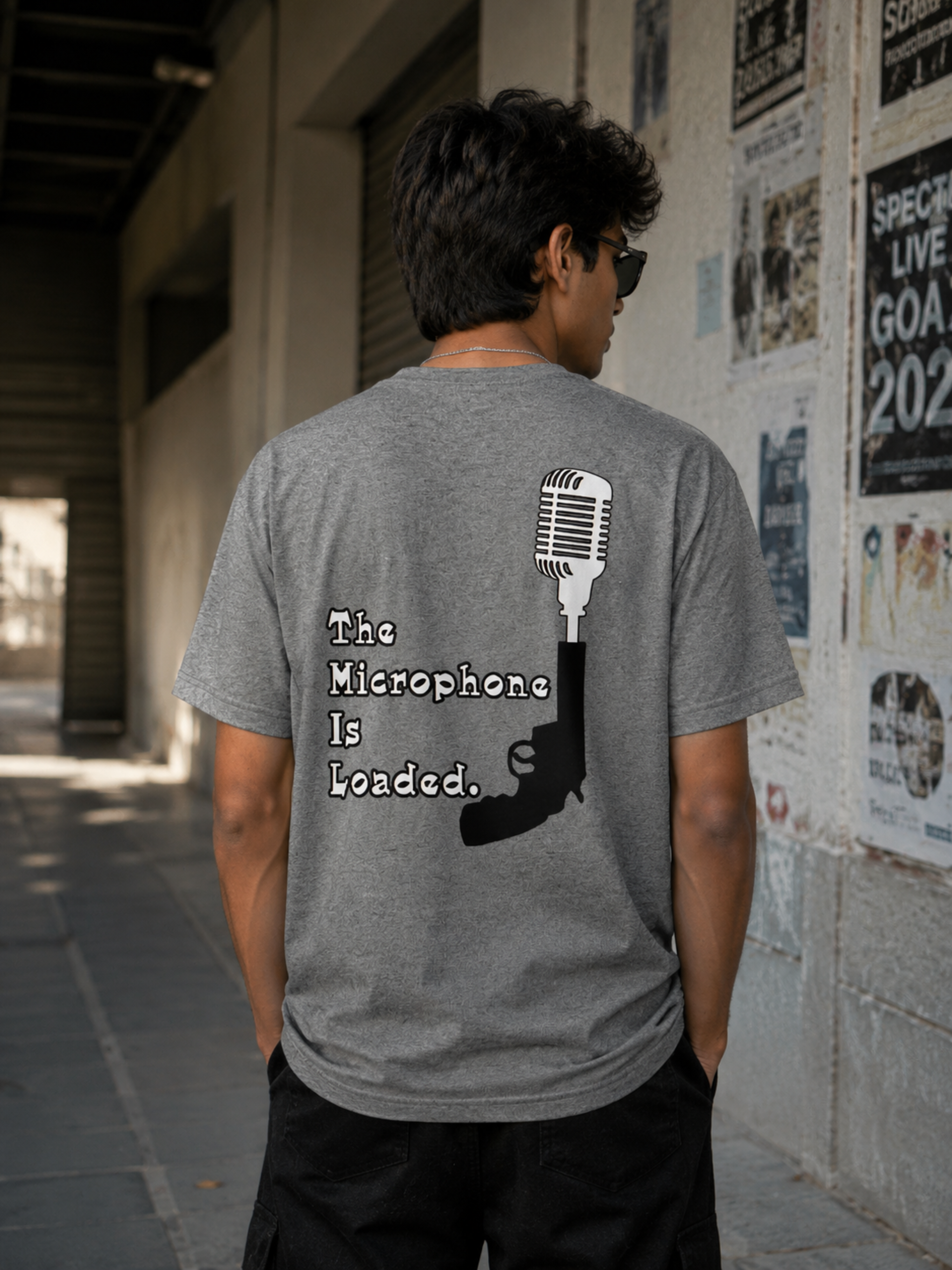 A man viewed from behind wearing a charcoal grey t-shirt with a black graphic of a microphone and gun and the text 'The Microphone Is Loaded.' in a street setting.