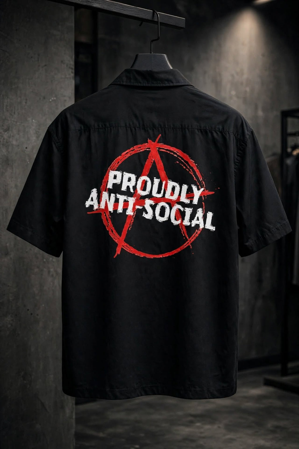 A black short-sleeve button-up shirt hanging on a hanger, showing the back with a large red anarchy symbol and white text reading 'PROUDLY ANTI-SOCIAL' on a dark indoor wall.