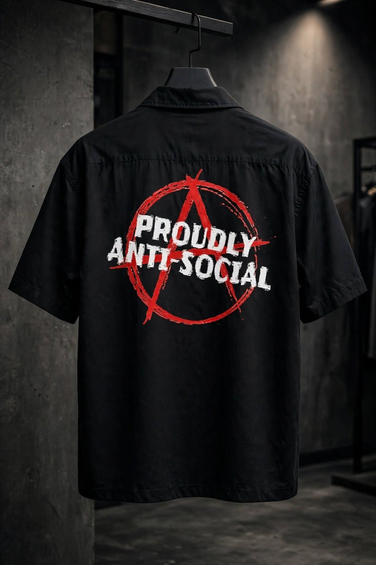 A black short-sleeve button-up shirt hanging on a hanger, showing the back with a large red anarchy symbol and white text reading 'PROUDLY ANTI-SOCIAL' on a dark indoor wall.