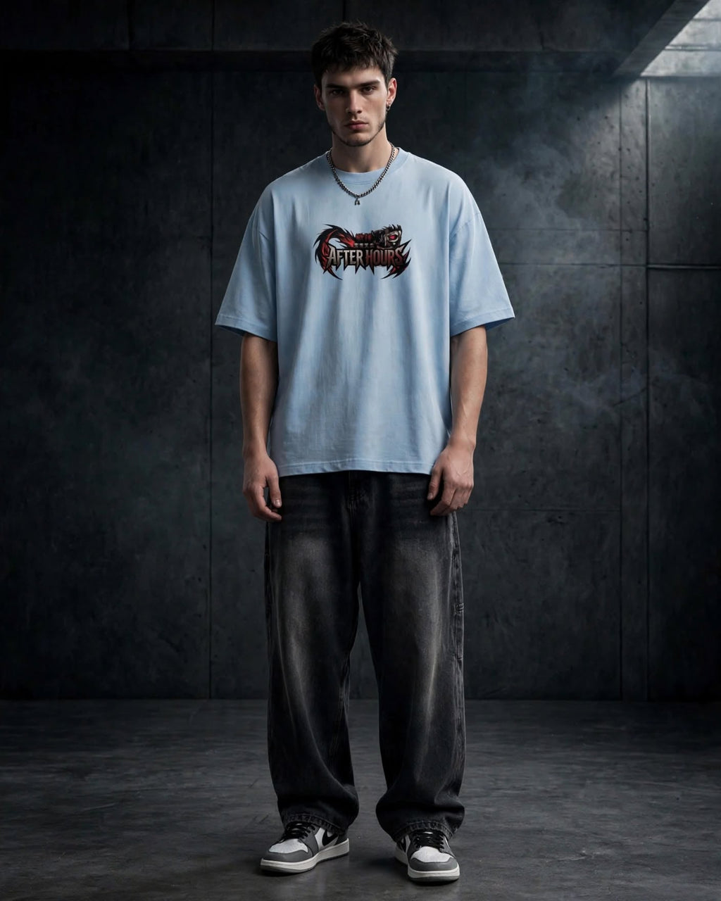 A man wearing a light blue t-shirt with a black and red graphic print on the chest, paired with dark grey baggy jeans and white sneakers, standing against a dark textured studio wall.