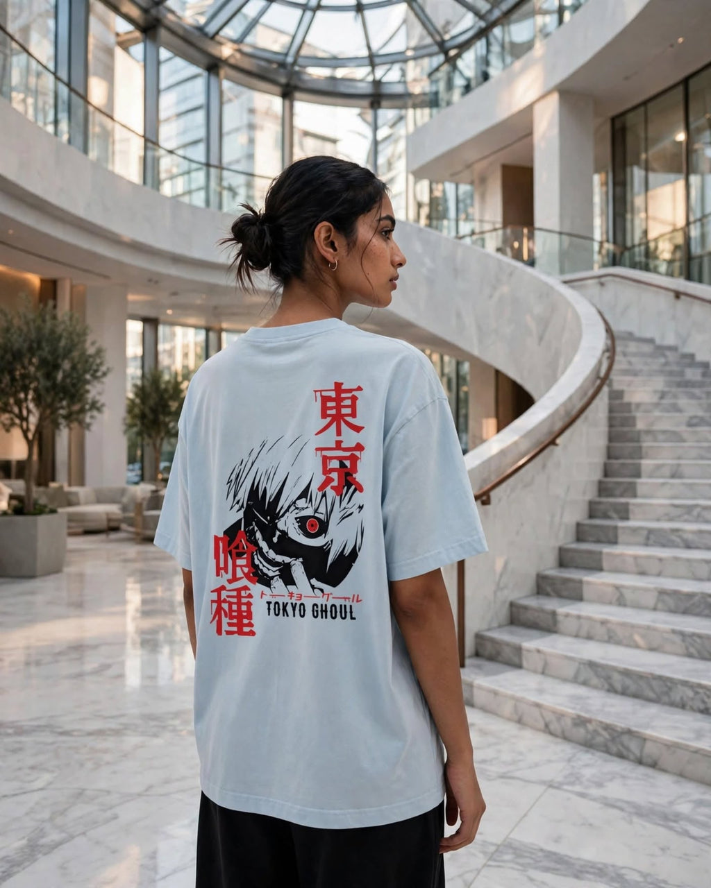 A woman stands in a grand, modern lobby with a marble staircase, wearing an oversized light blue T-shirt with a large back graphic featuring a black and white monster face, red Japanese characters, and 'TOKYO GHOUL' text.