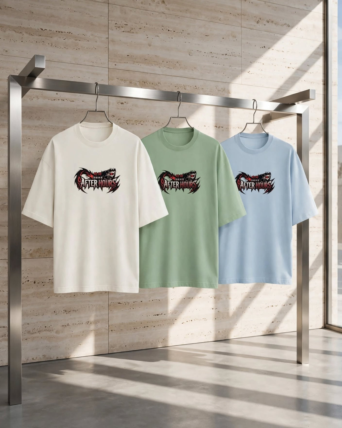 Three oversized short-sleeve T-shirts hang on a metal rack in a modern interior with warm sunlight and stone-textured walls. The shirts are off-white, light green, and light blue, each featuring a large black and red graphic print on the chest.