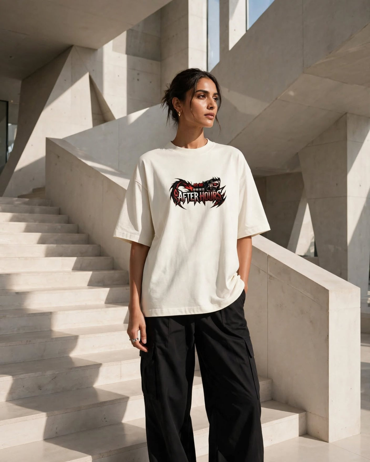 A woman wearing an oversized cream-colored t-shirt with a black and red graphic print that reads 'AFTER HOURS', paired with black pants, standing on concrete stairs in a modern architectural setting.