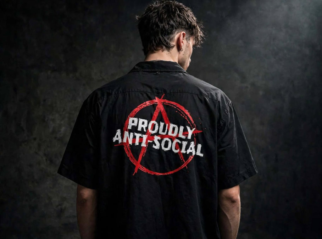 A man viewed from behind wearing a black short-sleeve button-up shirt with a red anarchy symbol and the words 'PROUDLY ANTI-SOCIAL' printed across the back, set against a dark textured studio background.