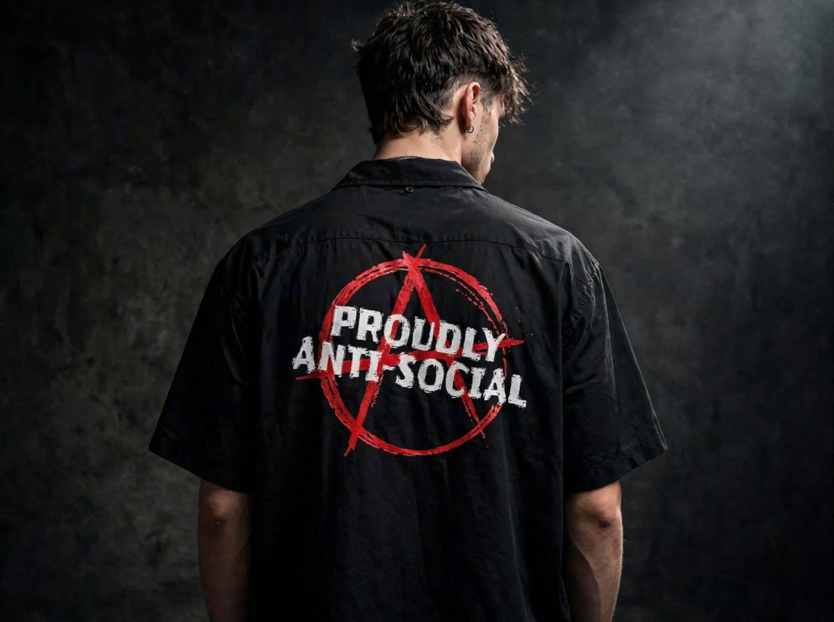 A man viewed from behind wearing a black short-sleeve button-up shirt with a red anarchy symbol and the words 'PROUDLY ANTI-SOCIAL' printed across the back, set against a dark textured studio background.