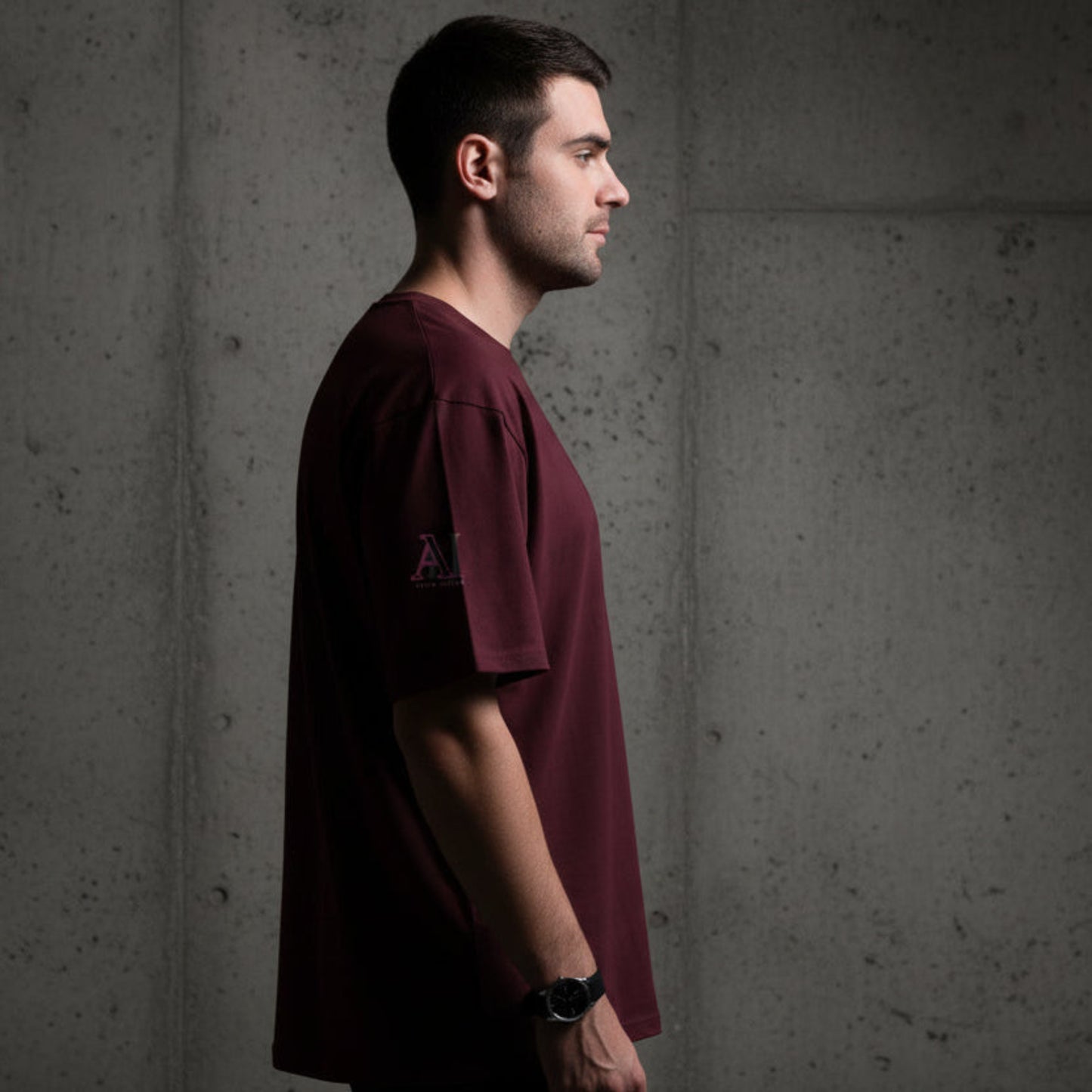 Man wearing a maroon t-shirt with a logo on the sleeve against a grey textured wall background