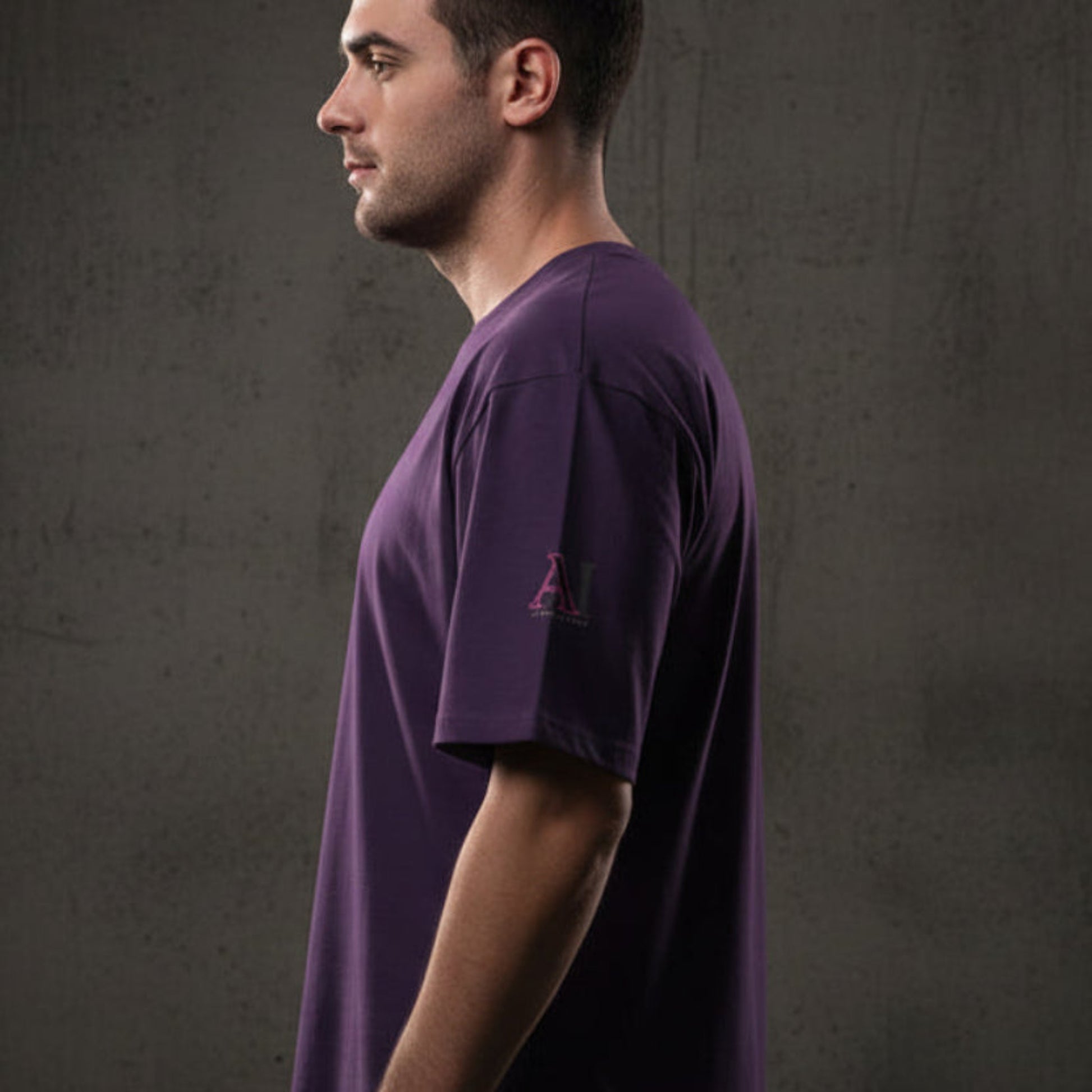 Man wearing a purple t-shirt with a logo on the sleeve against a grey textured wall background