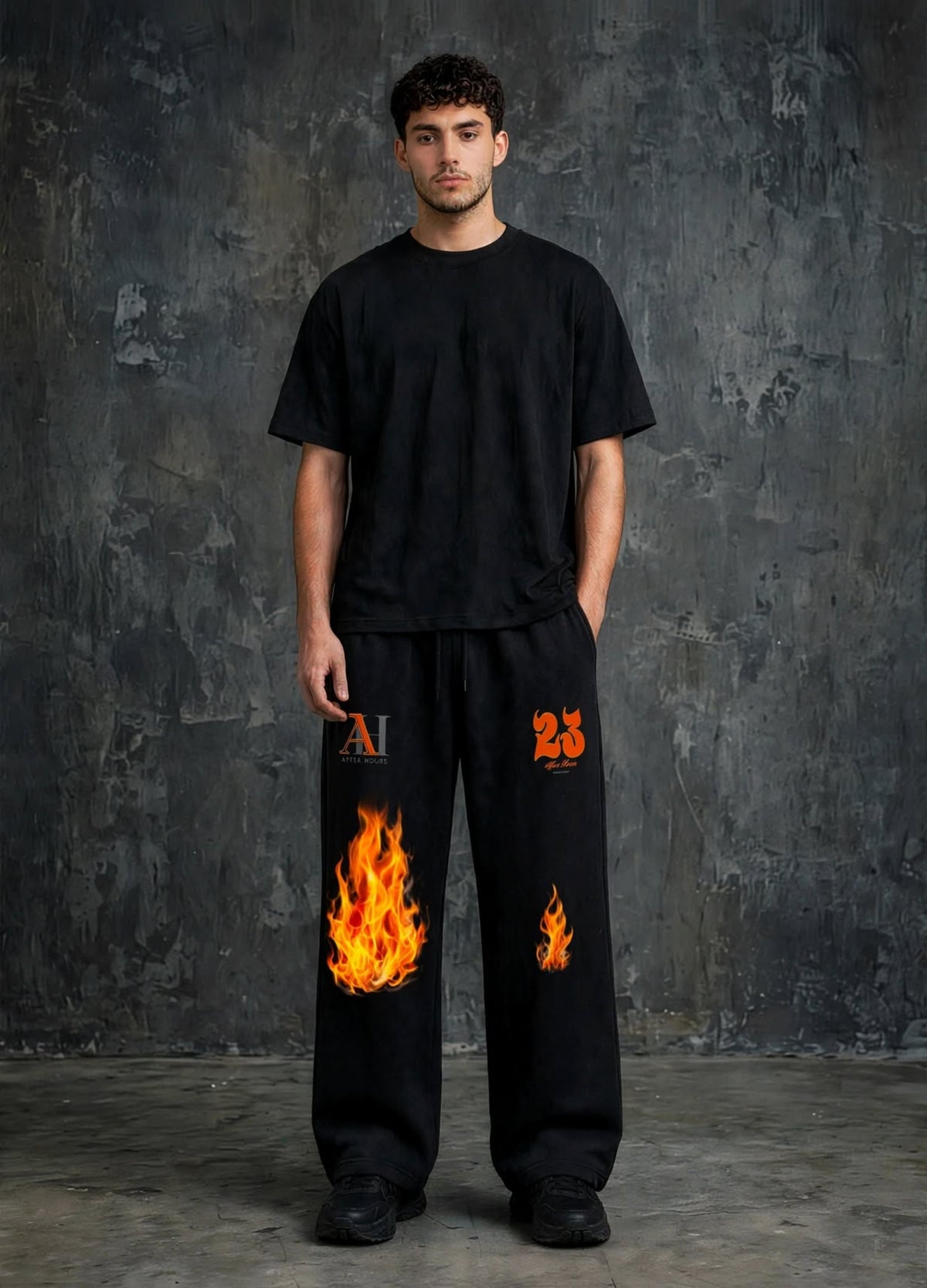 A man stands against a dark textured studio wall wearing black sweatpants with flame graphics and orange logo details, paired with a black t-shirt.