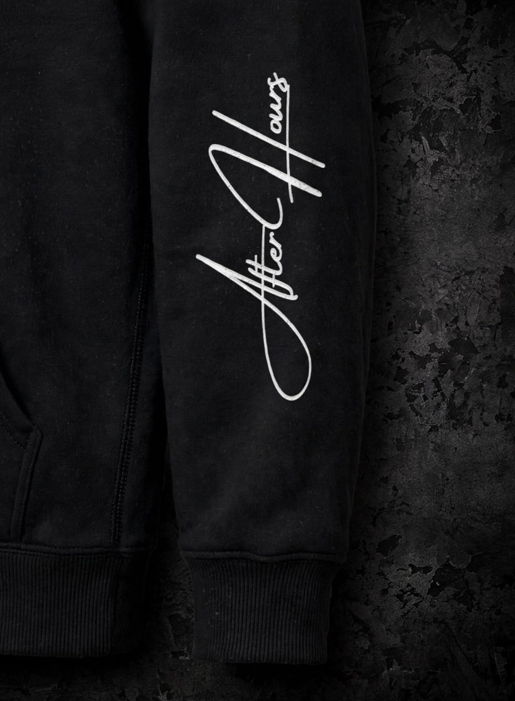 Close-up of a black hoodie sleeve featuring a large white cursive 'After Hours' graphic print, set against a dark textured background.