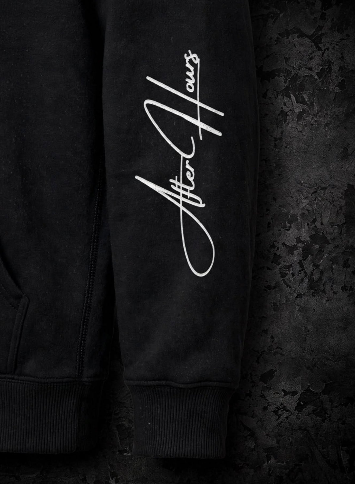 Close-up of a black hoodie sleeve featuring a large white cursive 'After Hours' graphic print, set against a dark textured background.