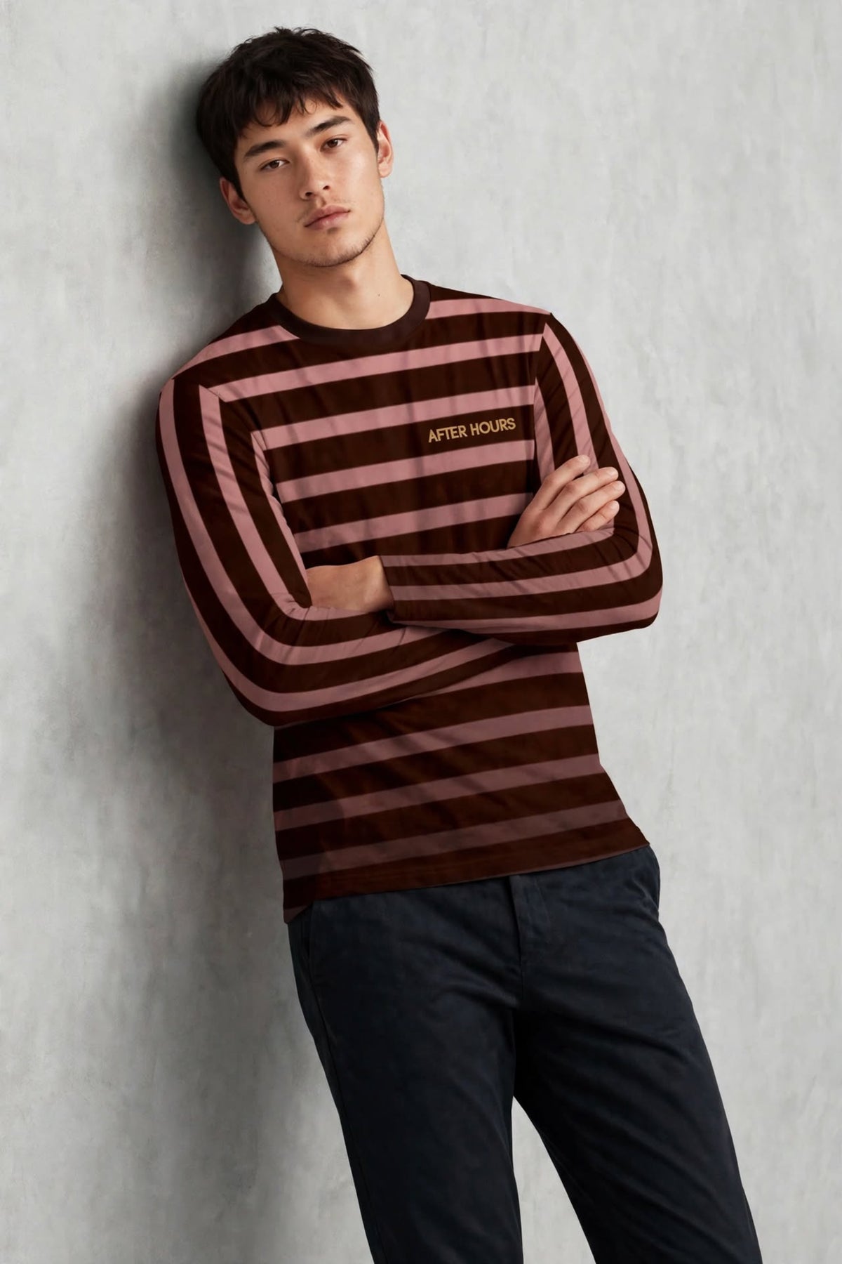 A man wearing a long-sleeve striped shirt with brown and dusty rose horizontal stripes, leaning against a textured grey wall. The shirt features the brand name 'AFTER HOURS' printed in gold on the left chest.