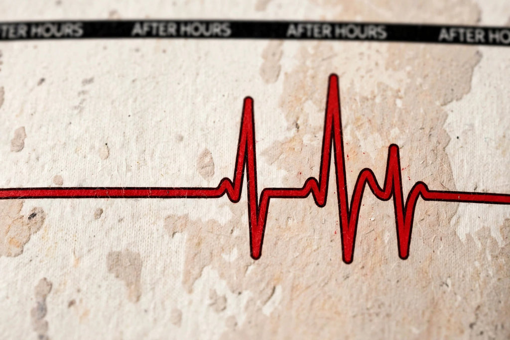 A close-up of a distressed beige sweatshirt featuring a red EKG heartbeat line with black labels reading 'AFTER HOURS' at the top.