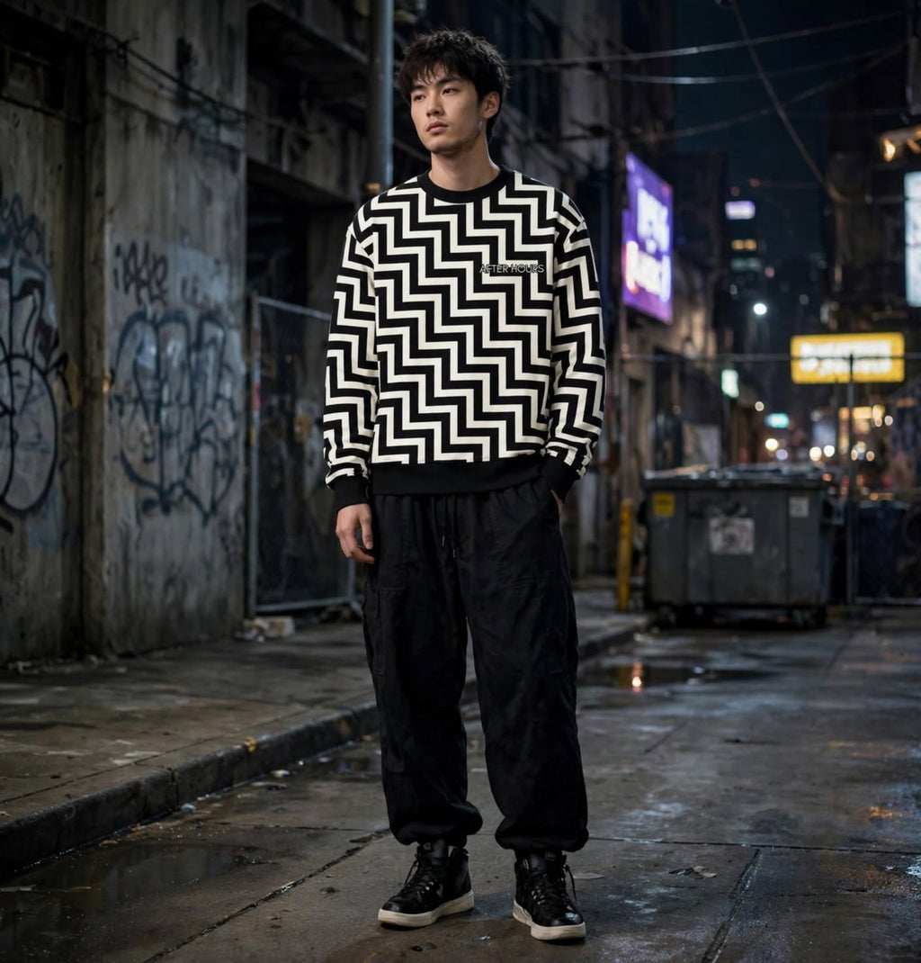 A man stands on a wet city street at night wearing a black and white geometric zigzag patterned sweater, black sweatpants, and black sneakers.