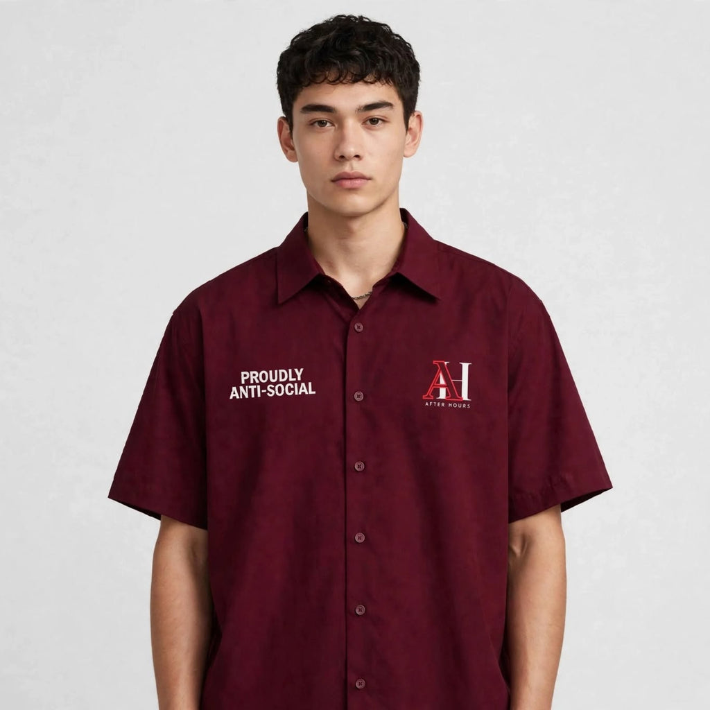 A young man wearing a short-sleeve button-up shirt in deep maroon. The shirt features white text on the right chest reading 'PROUDLY ANTI-SOCIAL' and a red and white 'AH' monogram logo on the left chest.