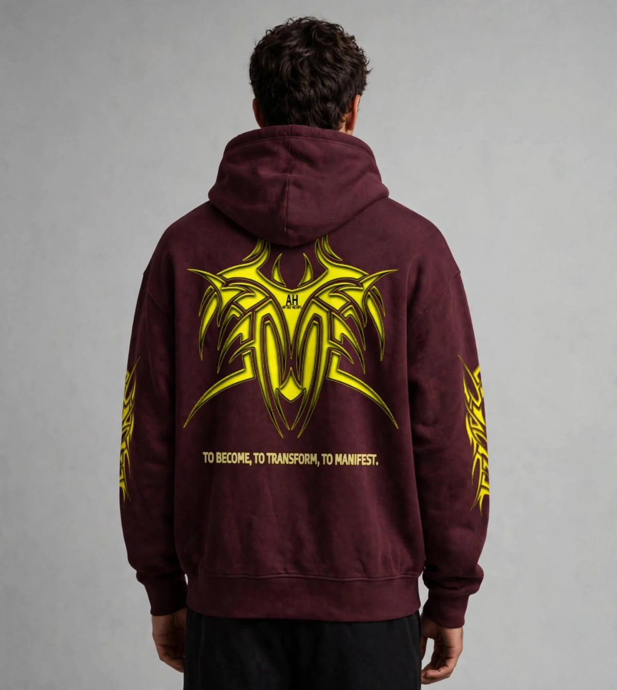 A man viewed from behind wearing a maroon hoodie with a large yellow graphic and text on the back, paired with black pants, against a neutral grey studio background.