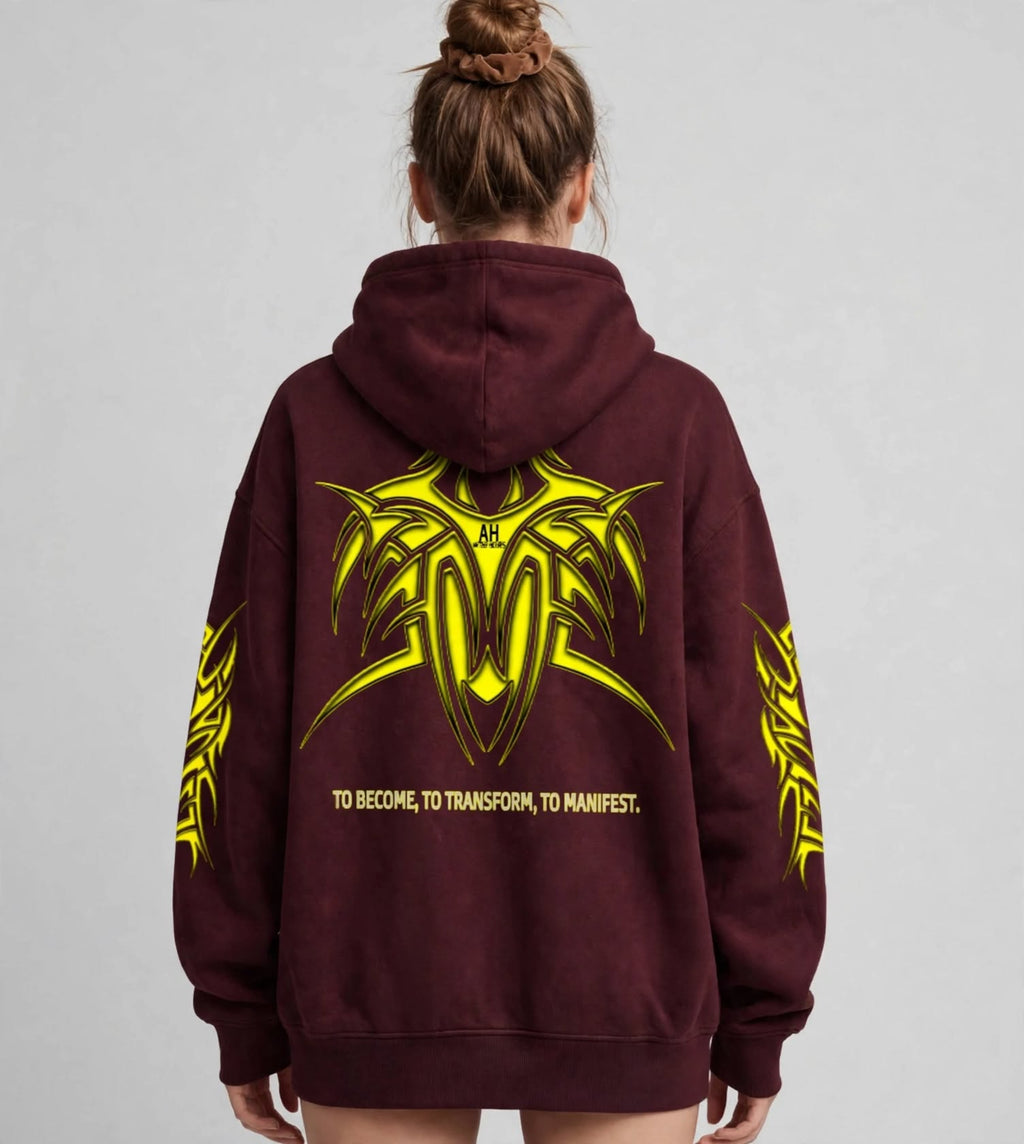 A person viewed from behind wearing an oversized maroon hoodie with a large yellow tribal-style graphic and text printed on the back, against a light grey studio background.