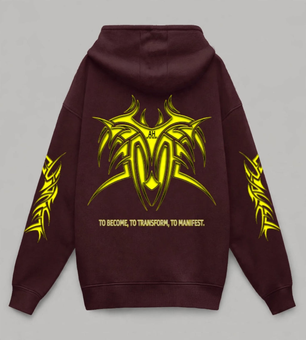 A dark maroon hoodie shown from the back, featuring a large yellow tribal graphic with the initials 'AH' in the center, the phrase 'TO BECOME, TO TRANSFORM, TO MANIFEST.' below it, and matching yellow graphics on both sleeves, set against a solid grey background.